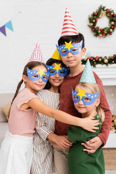 Positive interracial kids in party caps and masks hugging and looking at camera at home — Stock Photo