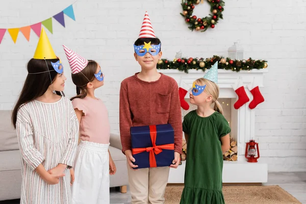 Asian boy holding present near friends in party caps and masks at home — Stock Photo