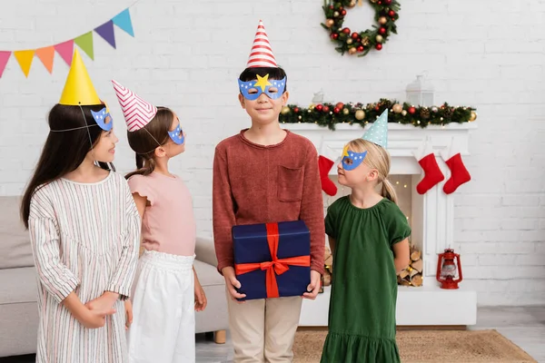 Asiatique garçon tenant cadeau près des amis dans les casquettes de fête et masques à la maison — Photo de stock