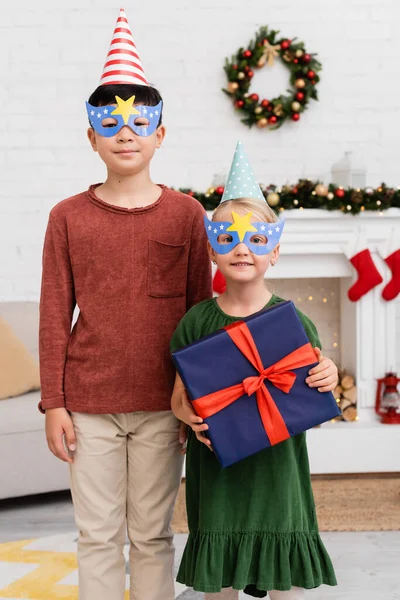 Multiethnic friends in party caps and masks holding present during birthday celebration in winter — Stock Photo