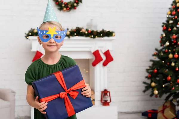 Ragazza sorridente in berretto partito e maschera in possesso di regalo con fiocco durante la festa di compleanno in inverno — Foto stock