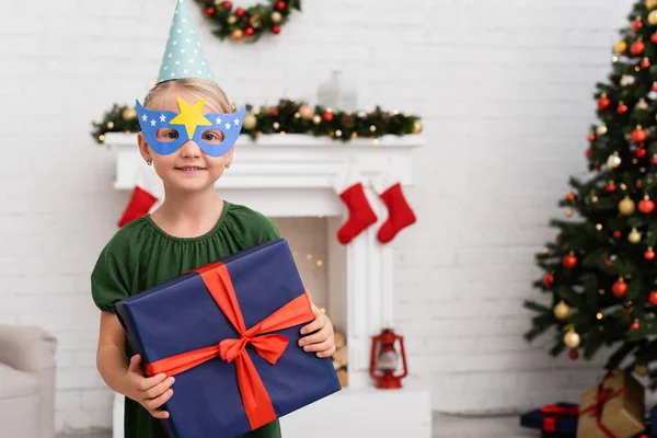 Smiling girl in party cap and mask holding gift with bow during birthday celebration in winter — Stock Photo