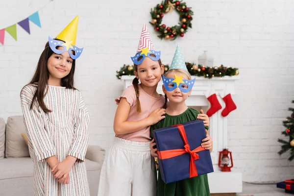 Smiling girls in party caps and masks hugging friend with present near christmas decor at home — Stock Photo