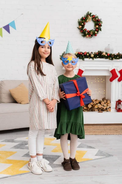 Smiling girls in party caps and masks holding present during birthday celebration in living room — Stock Photo