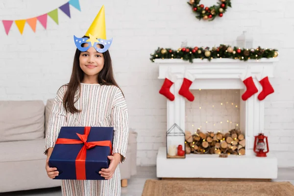 Smiling girl in party mask and cap holding gift with ribbon during birthday celebration in winter at home — Stock Photo