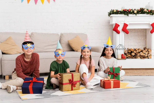 Улыбающиеся мультиэтнические дети в шапочках для вечеринок и масках, смотрящие в камеру возле подарков во время дня рождения дома — стоковое фото