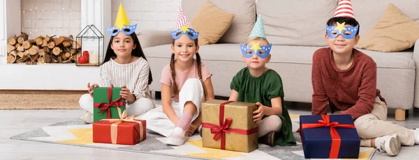Позитивные мультиэтнические дети в шапочках для вечеринок и масках, смотрящие в камеру возле подарков дома, баннер — стоковое фото