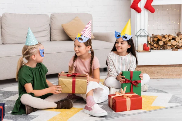 Positivo meninas segurando presentes perto de amigo em boné de festa e máscara em casa — Fotografia de Stock