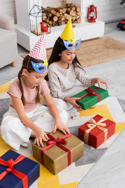 Vista de ángulo alto de las niñas preadolescentes en gorras de fiesta y máscaras mirando los regalos durante el cumpleaños en casa - foto de stock