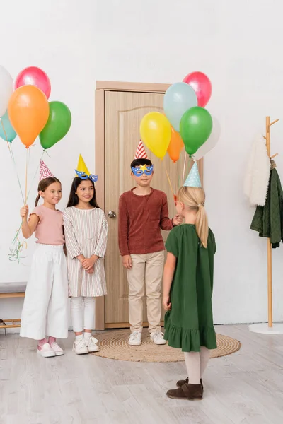 Interracial kids in party masks and caps holding balloons near friend in hallway — Stock Photo