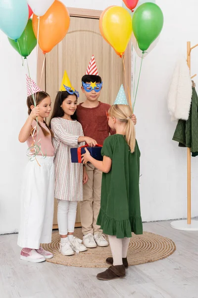 Interracial children in party caps holding balloons and present near friend in hallway at home — Stock Photo