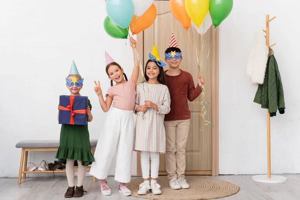 Niños alegres multiétnicos en gorras de fiesta sosteniendo globos y regalos mientras miran a la cámara en el pasillo en casa - foto de stock