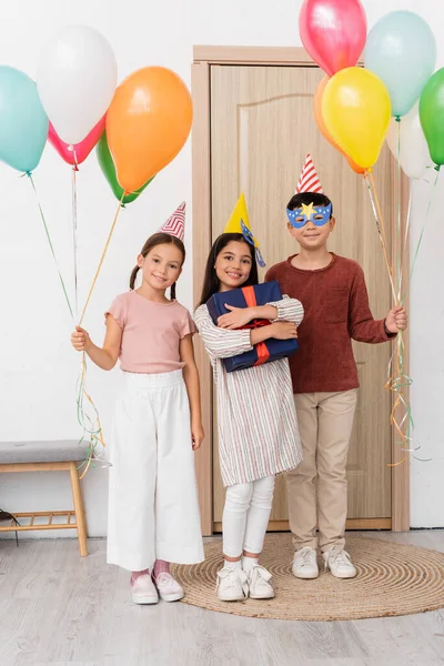 Sorrindo menina na tampa do partido segurando caixa de presente perto de amigos inter-raciais com balões no corredor em casa — Fotografia de Stock