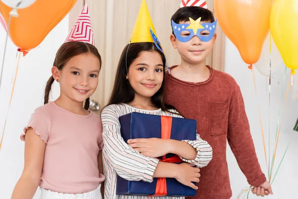 Smiling interracial kids in party caps holding gift and balloons in hallway at home — Stock Photo