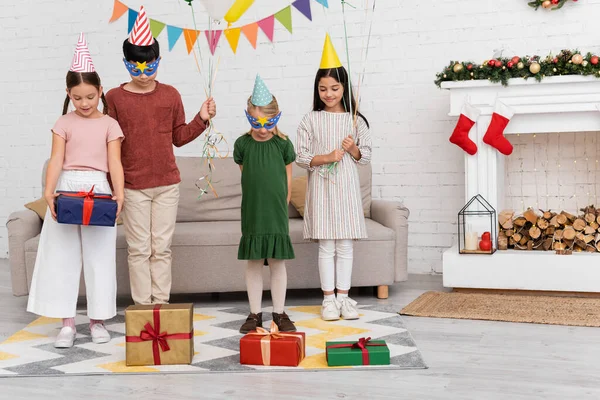 Sonrientes niños multiétnicos en gorras de fiesta sosteniendo globos cerca de regalos y decoración de Navidad en casa - foto de stock