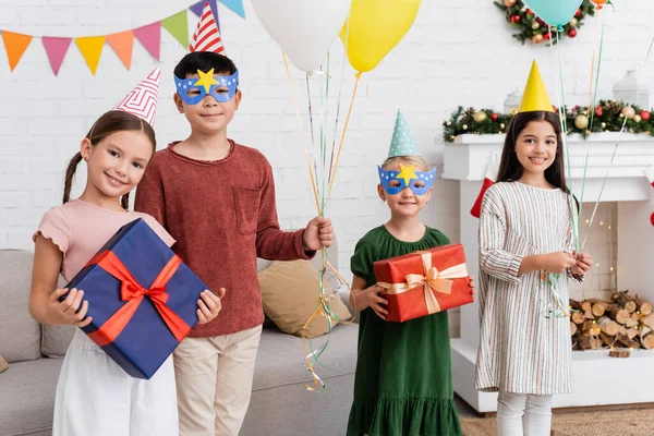 Niños multiétnicos en gorras de fiesta sosteniendo globos y regalos durante la celebración del cumpleaños en invierno en casa - foto de stock