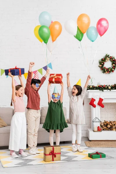 Emocionados niños multiétnicos en gorras de fiesta y máscaras sosteniendo globos cerca de amigos con regalo de cumpleaños y decoración de Navidad en casa - foto de stock