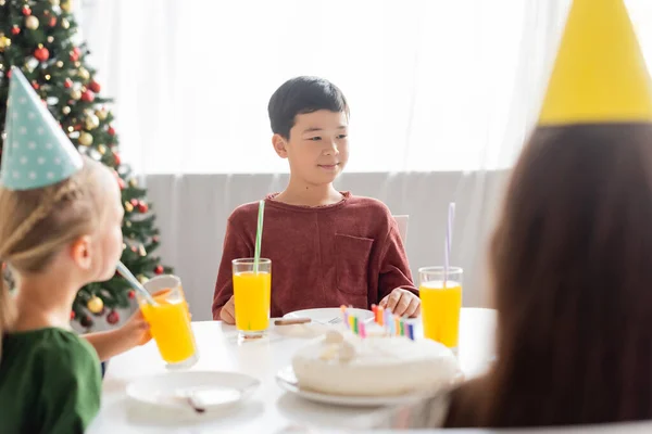 Asian kid sitting near blurred friends in party caps and birthday cake at home in winter — Stock Photo