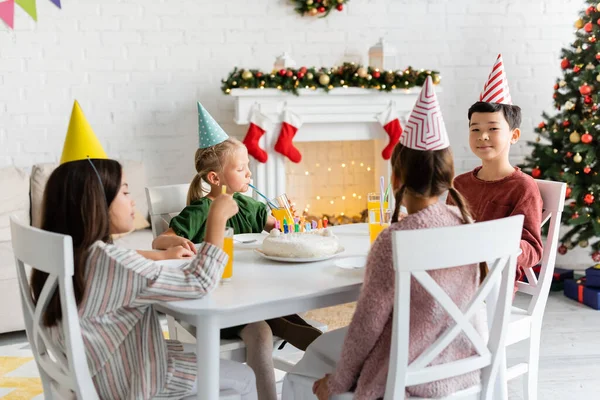 Interracial kids in party caps sitting near birthday cake and orange juice at home in winter — Stock Photo