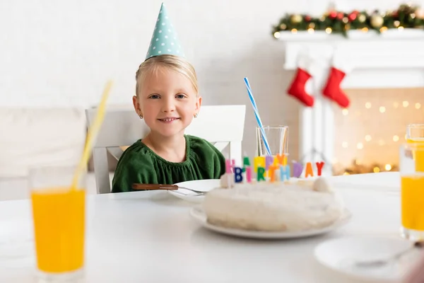 Chica sonriente en gorra de fiesta mirando a la cámara cerca de pastel de cumpleaños y jugo de naranja en casa - foto de stock