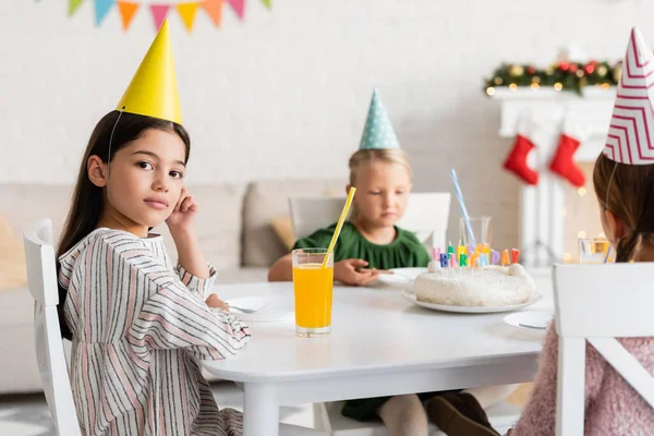Ragazza in berretto partito guardando la fotocamera vicino agli amici e torta di compleanno a casa — Foto stock