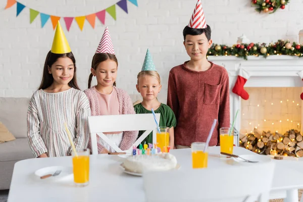 Sonriendo niños interracial en gorras de fiesta mirando pastel de cumpleaños durante la fiesta en casa en invierno - foto de stock