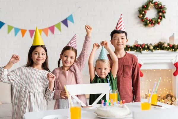 Excited multiethnic children in party caps looking at camera near birthday cake during party at home in winter — Stock Photo