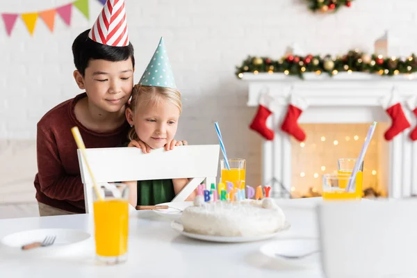 Niños multiétnicos en gorras de fiesta mirando pastel de cumpleaños con velas y jugo de naranja en casa - foto de stock