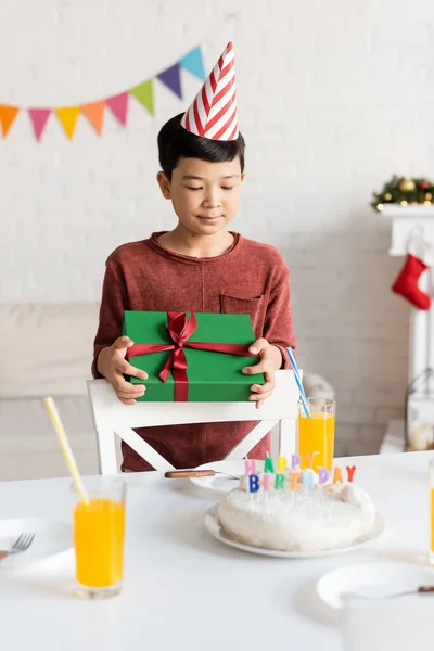 Asiatischer Junge mit Partymütze hält Geschenk in der Nähe von Geburtstagstorte und Orangensaft zu Hause — Stockfoto