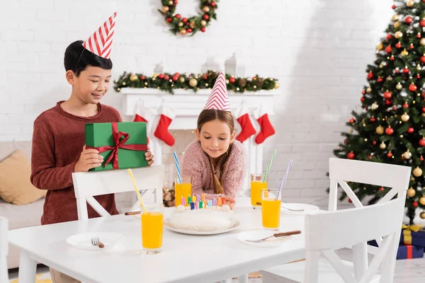 Улыбающийся азиатский мальчик в шапочке для вечеринок держит подарок рядом с другом и торт на день рождения в доме зимой — Stock Photo