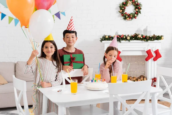 Enfants multiethniques tenant des ballons et présents près du gâteau d'anniversaire à la maison en hiver — Photo de stock