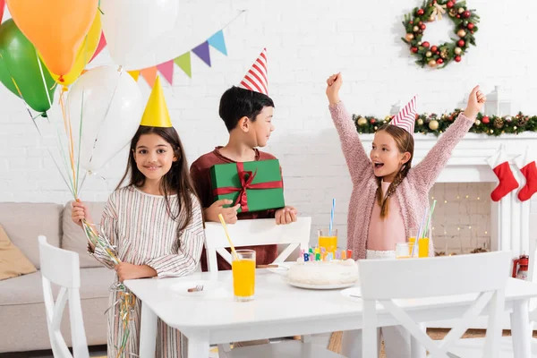 Smiling multiethnic kids in party caps holding balloons and gift near excited friend and birthday cake at home in winter — Stock Photo