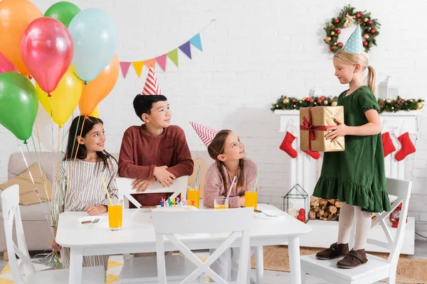 Межрасовые дети в шапочках для вечеринок держат воздушные шары рядом с другом с подарком и праздничным тортом дома — стоковое фото