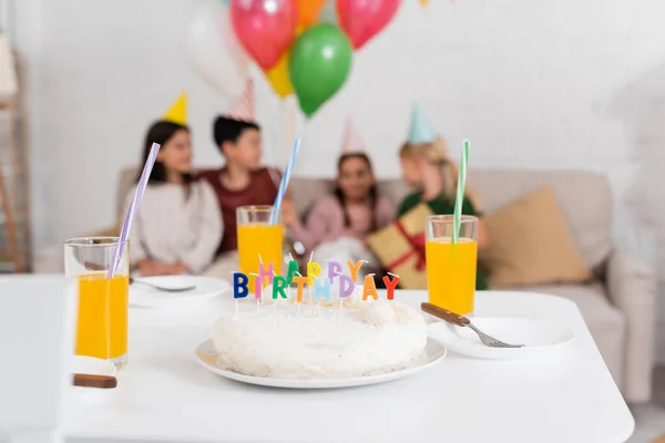 Birthday cake and orange juice on table near blurred kids at home — Stock Photo