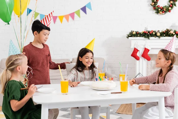 Happy girls in party caps looking at birthday cake near asian friend with balloons at home — Stock Photo