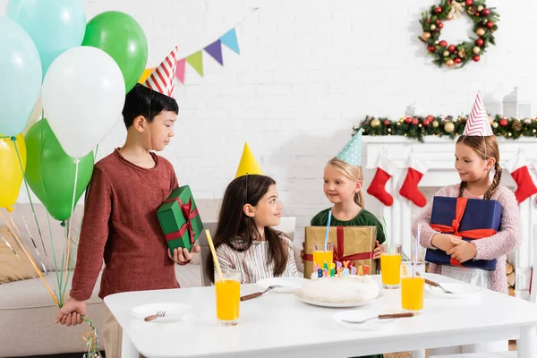 Niños multiétnicos en gorras de fiesta sosteniendo regalos y globos cerca de amigos y pastel de cumpleaños en casa - foto de stock