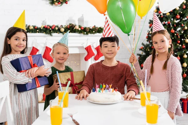 Meninas sorridentes em bonés de festa segurando presentes e balões perto de amigo asiático e bolo de aniversário em casa no inverno — Fotografia de Stock