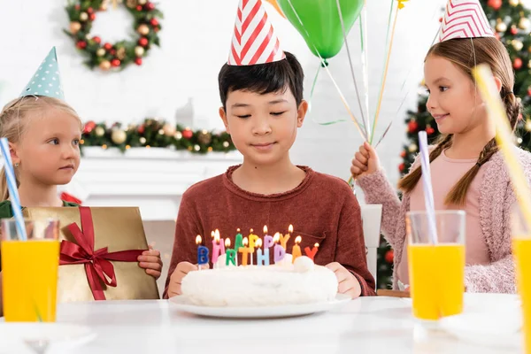 Asiático chico en partido gorra mirando cumpleaños pastel con velas cerca de amigos con regalo y globos en casa - foto de stock