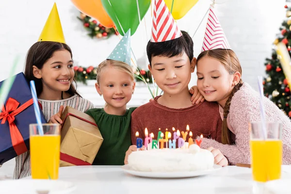 Multiethnic kids in party caps holding presents near birthday cake and orange juice at home — Stock Photo