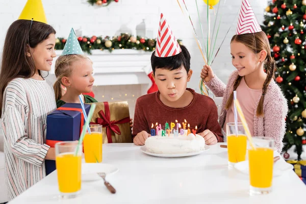 Asiático chico en partido gorra soplando fuera velas en cumpleaños pastel cerca amigos con presente en casa - foto de stock