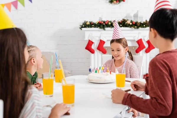 Lächelndes Mädchen mit Partymütze schaut Geburtstagstorte neben verschwommenen Freunden zu Hause an — Stockfoto