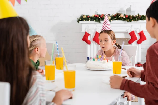 Chica en gorra de fiesta soplando velas en pastel de cumpleaños cerca de amigos y jugo de naranja en casa - foto de stock