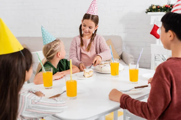 Chica sonriente en gorra de fiesta sirviendo pastel de cumpleaños a amigos borrosos cerca del jugo de naranja en casa - foto de stock