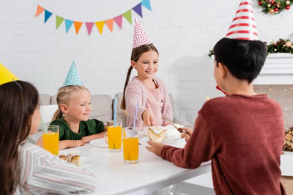 Kid in party cap giving birthday cake to blurred friend near children at home — Stock Photo