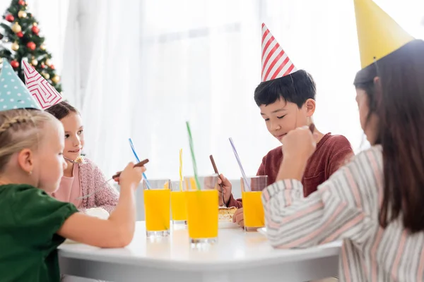 Asiático chico en partido gorra mirando cumpleaños pastel cerca amigos y naranja jugo en sala de estar - foto de stock