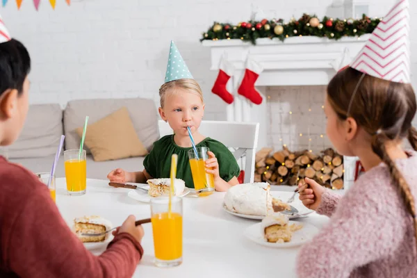 Niño con gorra de fiesta bebiendo jugo de naranja cerca de amigos borrosos y pastel de cumpleaños en casa en invierno - foto de stock