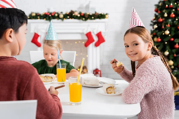 Chica sonriente en gorra de fiesta mirando a la cámara y sosteniendo pastel de cumpleaños cerca de amigos multiétnicos y jugo de naranja en casa en invierno - foto de stock