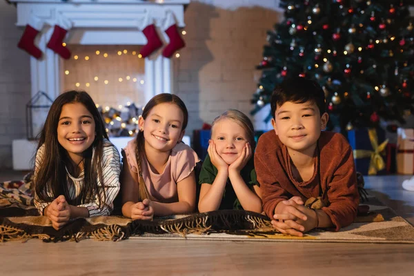 Lächelnde Kinder blicken in die Kamera, während sie auf einer Decke auf dem Boden neben dem Weihnachtsbaum liegen — Stockfoto