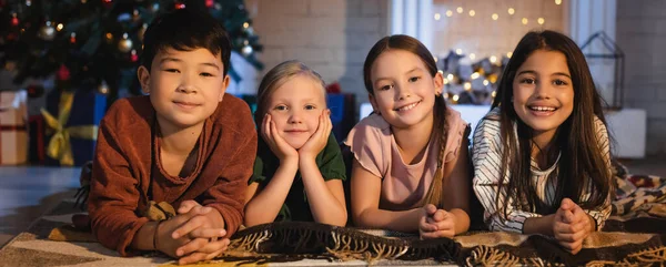 Niños multiétnicos sonriendo a la cámara cerca del borroso árbol de Navidad en casa por la noche, pancarta - foto de stock