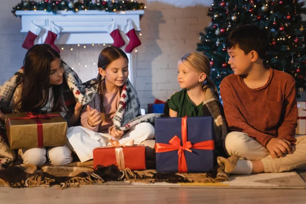 Preteen filles avec couverture tenant cadeau près d'amis multiethniques et arbre de Noël à la maison — Photo de stock